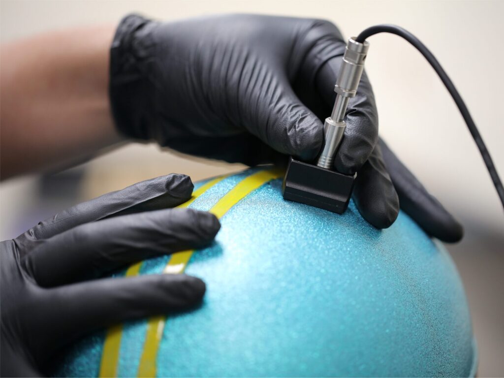 Technician uses an ultrasonic probe to measure the powder coating thickness of blue paint on a helmet.