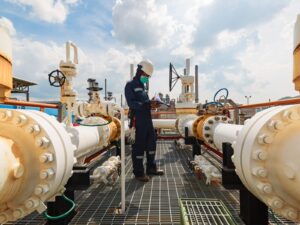 An expert performs a piping inspection at an oil and gas refinery. He holds a clipboard and wears a hardhat.