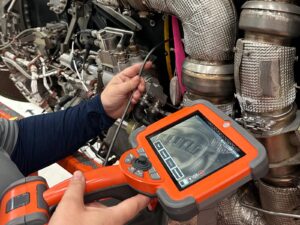 NDT technician guides a borescope cable into an airplane engine. She views the camera's footage on a handheld borescope screen.