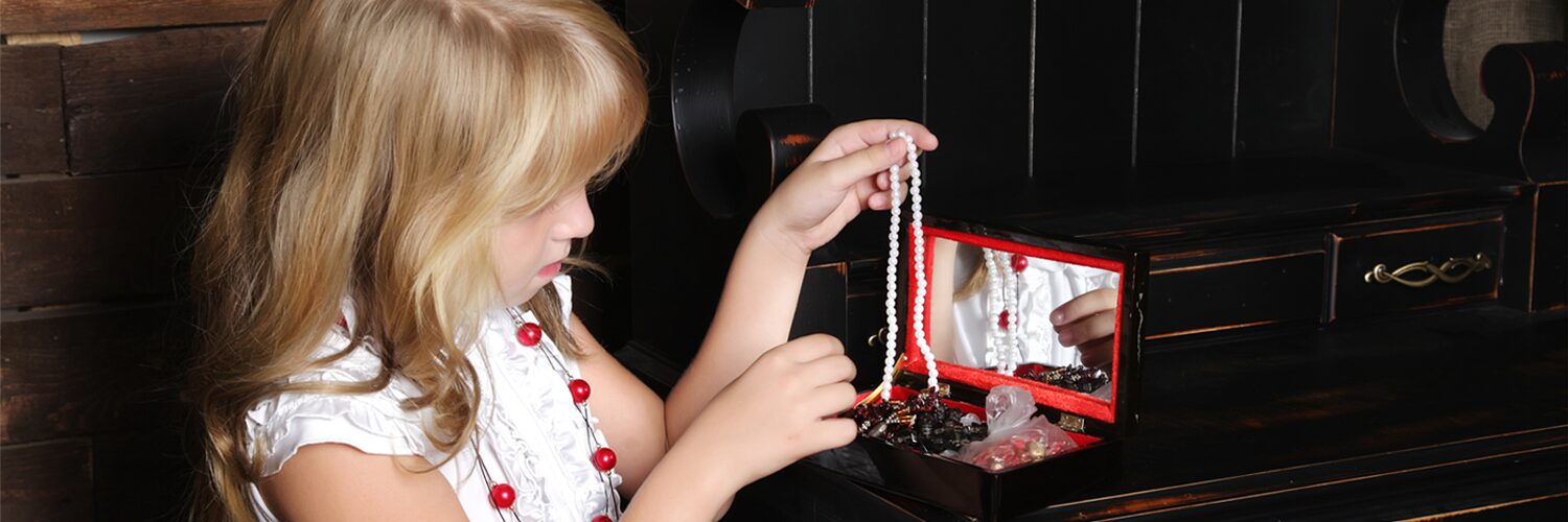 Little girl holds a beaded necklace up to a jewel case and mirror