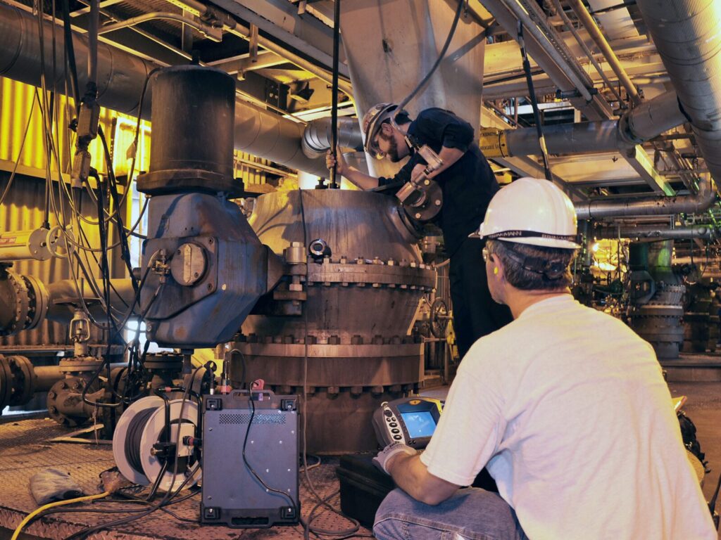 A field technician lowers a remotely operated camera into a tank as a technician with a monitoring screen observes.