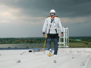 A rope access technician crosses the flat rooftop of a building. He wears a harness connected to a tie-back anchor on the roof via lanyard. In the distance, clouds gather.