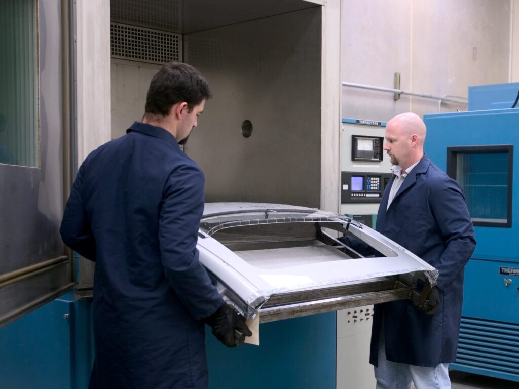 Two lab technicians load an automotive component into an environmental chamber for thermal testing.