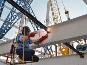 Industrial welder welds two steel beams together. The welder is suspended mid-air with a rope platform.