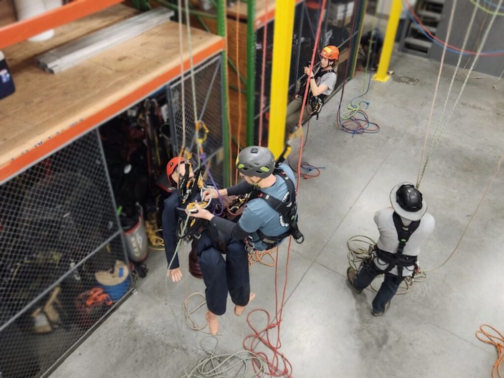 View of three rope access technicians from above. One suspended technician adjusts the harness on a mannequin. Below, a technician holds several rope lines.