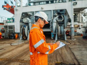 Inspector on a ship deck takes notes on a clipboard.