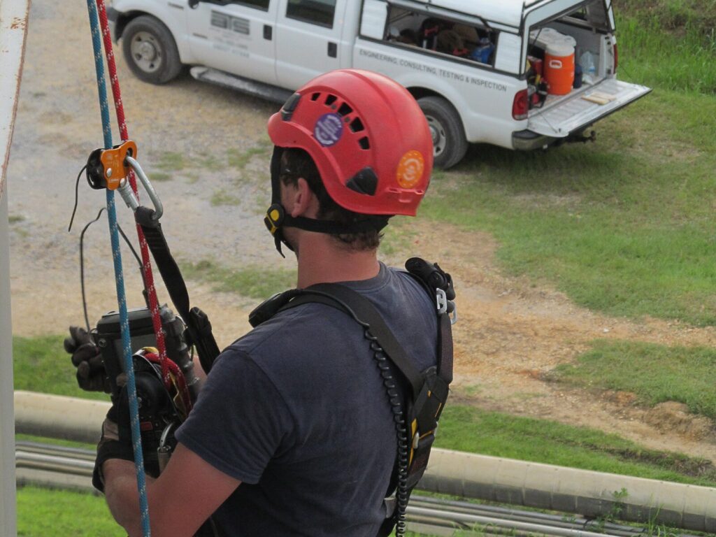 Rope access technician suspended from an aboveground tank looks down at a grassy lawn and ATS truck