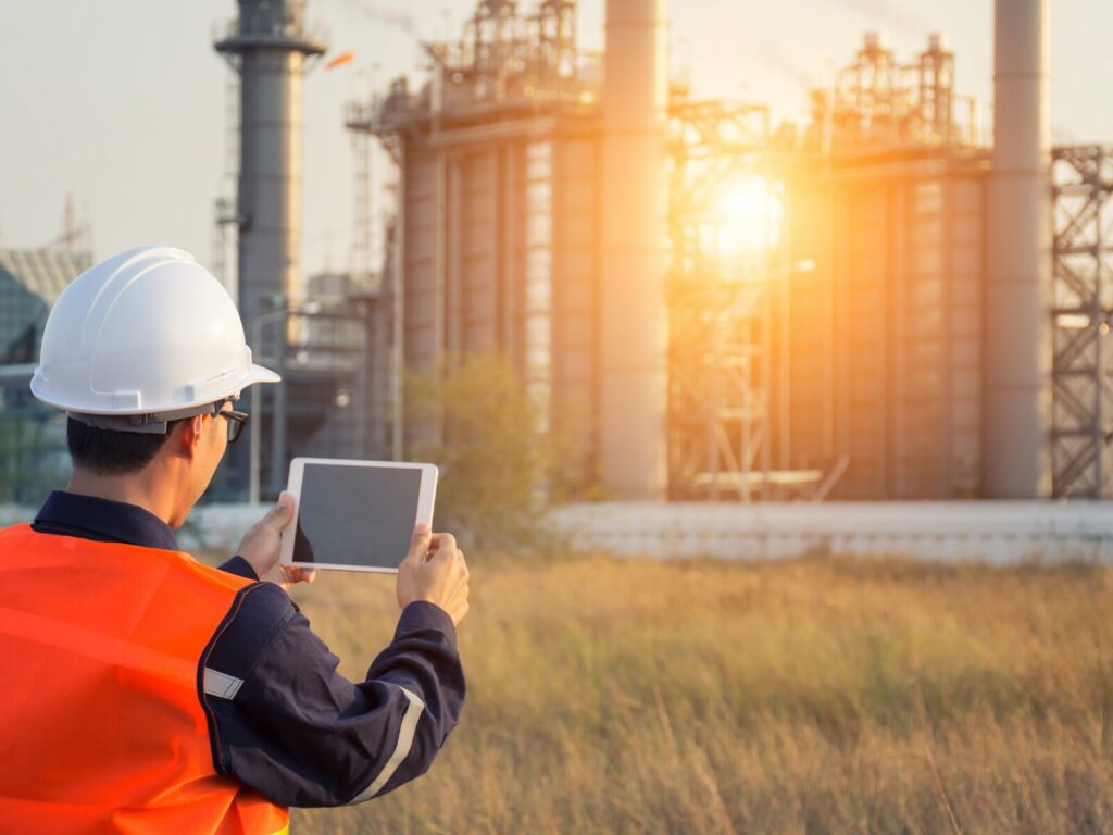 Technician holds up a tablet. An industrial processing plant is in the background.