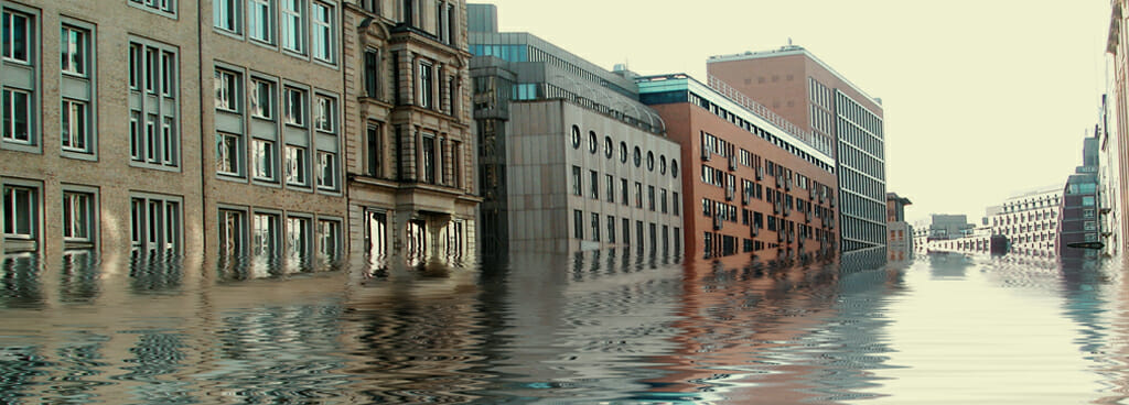 A flooded historic city street partially reflected in the rippling water. The flood damage is already starting to show along the high water lines under close inspection of the wet bricks.