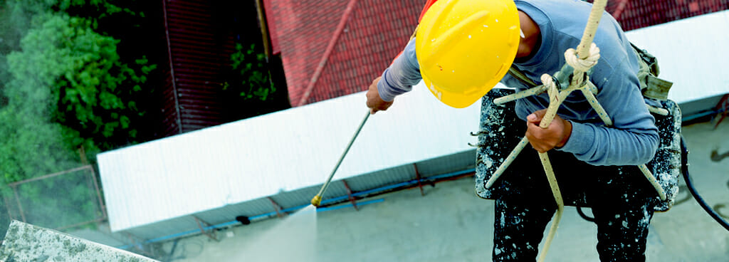 A man rigged to a safety rope rappels over an industrial building porch. He uses a hose to spray the porch. He wears a yellow hard hat and uses safety tools to show he provides building envelope consulting services.