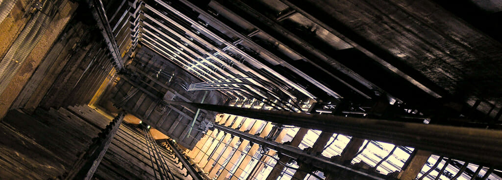 View down an elevator shaft as seen during equipment failure analysis after an elevator malfunction. The elevator is stuck in the dark at the bottom of the shaft. On the right side of the image, golden light filters through the rungs of the shaft.