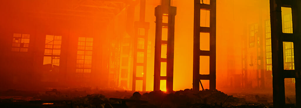 Inside a warehouse on fire. The flames and smoke glow orange in the distance. Ash covers the ground.