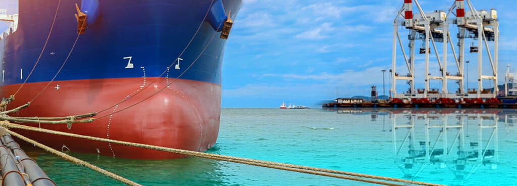 A ship's bow, partially submerged by marine water. The bow is dark blue with a red stripe for the bulbous bow that juts out into the brilliant green water. The ship is tethered to a port with massive ropes. On the other side of the harbor is a set of container cranes. A marine structural engineer works with all of these vessels and structures.