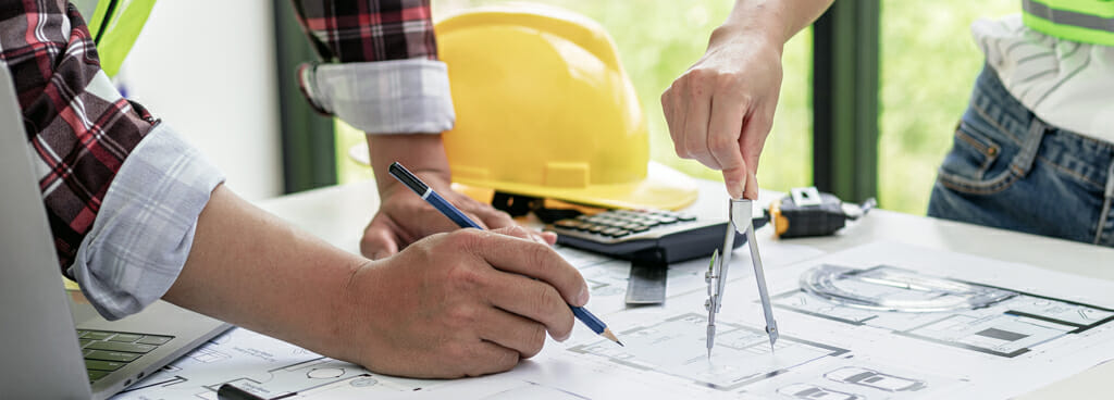As one of the structural design services provided by ATS, architect and structural engineer review the blueprint for a building. One holds a blue pencil, the other holds a compass. In the background, a yellow hardhat rests on the table.