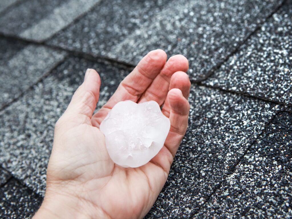 A hand holds a palm-sized hailstone by a roof