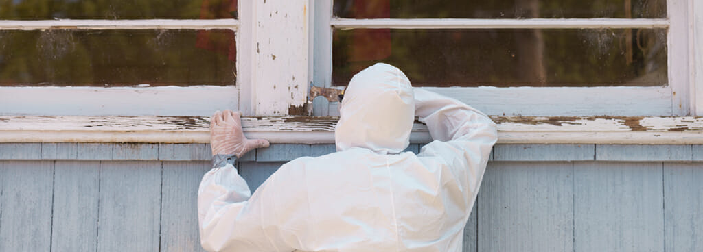 A lead abatement contractor inspects white and cracking lead-based paint on a window casing.