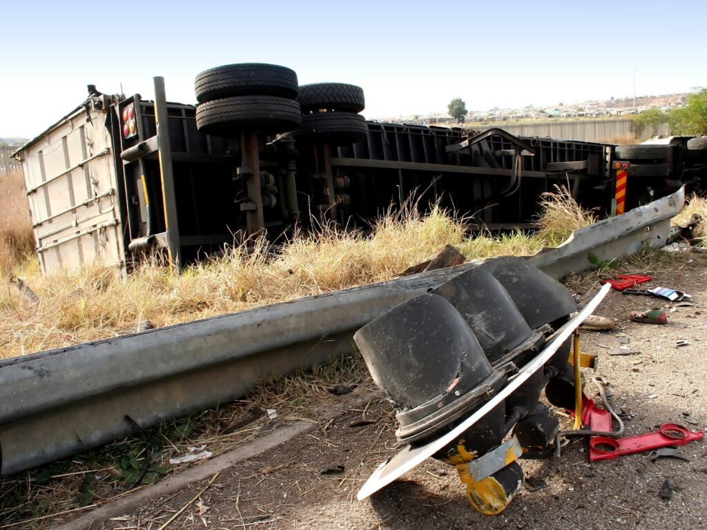 Highway guardrail and debris after an accident. In the background, an overturned eight-wheel truck.