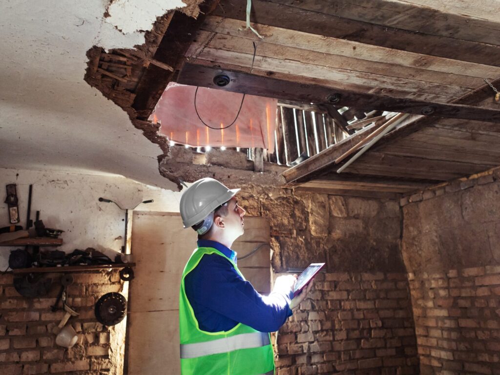 Engineer inspects a massive hole in the ceiling of a building in disrepair. He wears protective clothing, a hard hat, and carries a tablet.