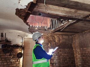 Engineer inspects a massive hole in the ceiling of a building in disrepair. He wears protective clothing, a hard hat, and carries a tablet.