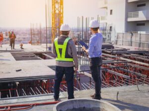 Two engineers stand at the edge of a construction site. The complex rebar system for a foundation is visible beyond them.