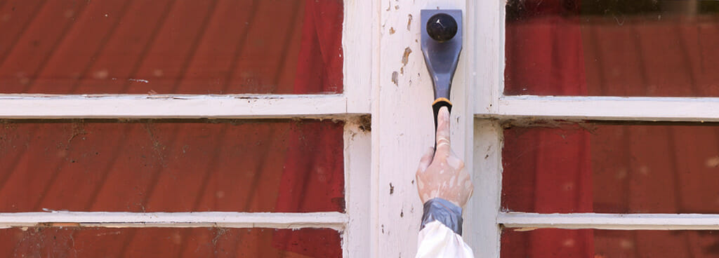 An EPA certified lead abatement contractor uses a trowel to scrape a sample of lead paint off the side of an old house.