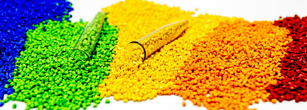 A collection of plastic pellets sorted by color to resemble a rainbow. Two test tubes sit atop the green and yellow piles to take samples for polymer composition analysis.