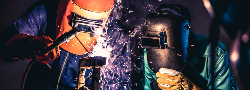 A welder uses a torch to weld a flat plate in a 1G weld test. A second welder watches the torch with interest. The welders wear orange and black helmets and thick gloves to protect them.
