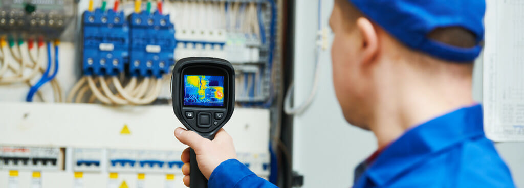 An inspector with infrared thermography certification uses a thermal imaging scanner to detect unusual heat signatures in an electrical panel.