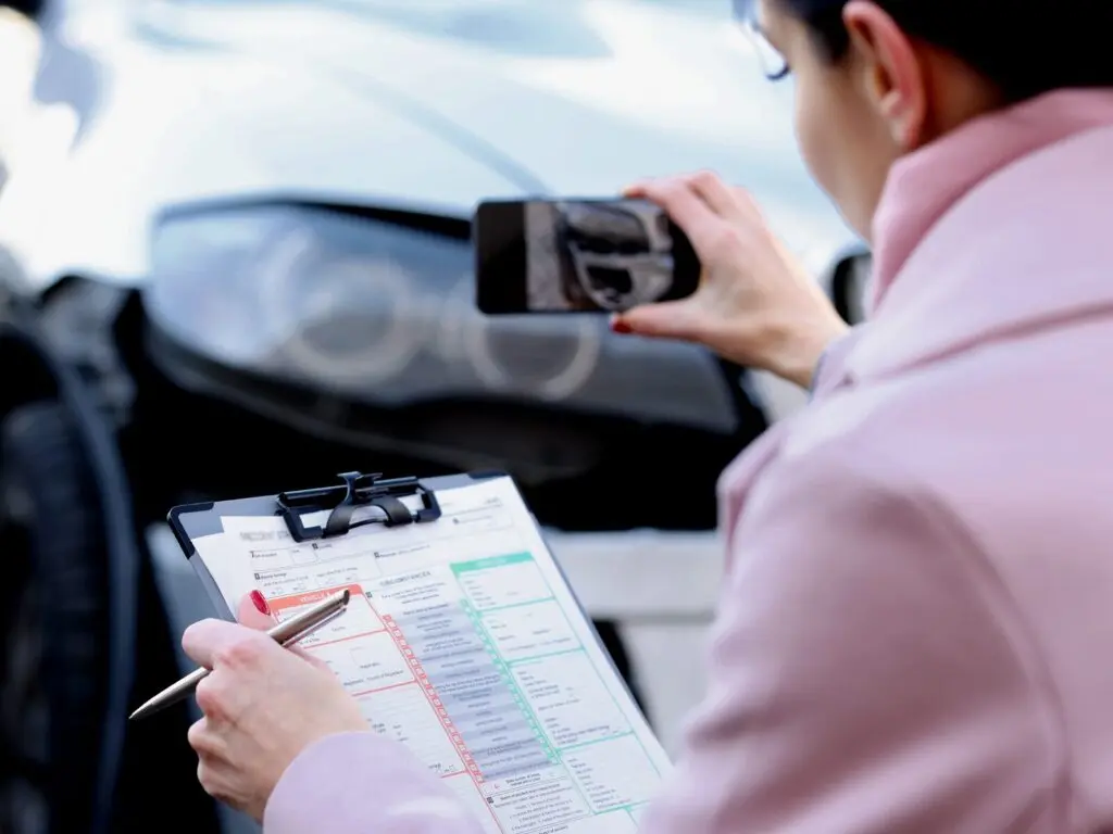 Woman takes a photo of a vehicle collision with her phone. She holds a clipboard and pen in her other hand.