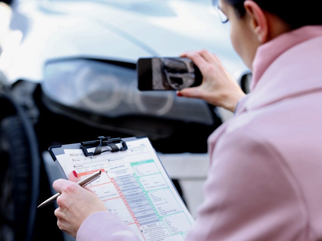 Woman takes a photo of a vehicle collision with her phone. She holds a clipboard and pen in her other hand.