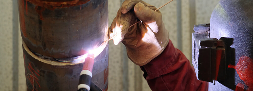 A welder reaches up to weld two large pipe pieces for a 6G Welding Certification test. They wear thick gloves and a jacket to protect their skin and a welding mask to protect their face and eyes. One gloved hand holds an electrode wire and the other a welding torch.