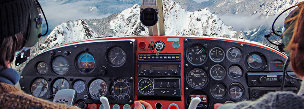 The flight instrument panel of a small aircraft flying over snowy mountains. The shoulders of the male pilot and female co-pilot are visible. The pilot wears a brown jacket. The co-pilot has short red hair. There are three rows of gauges in front of each of them. The anemometers are maintained with air velocity meter calibration.