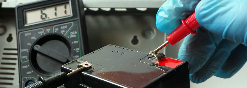 A technician handles a valve-regulated lead-acid battery during battery performance testing. In the background, a battery tester displays the voltage of the battery under test.
