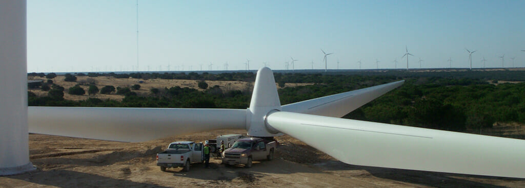 Two pick-up trucks are overshadowed by the massive blades of a downed wind turbine. Beyond the turbine is a desert landscape of bushes, clay earth, and a distant wind farm.