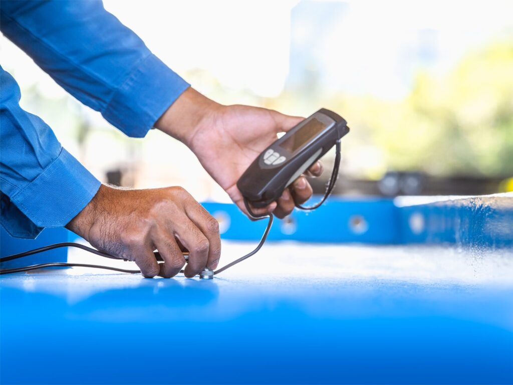 Technician uses a PosiTector to measure the thickness and gloss of blue paint on a pipeline