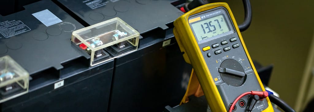 A technician uses a Fluke 87V digital multimeter to measure performance of a large car battery. The meter is grey with a protective yellow case.