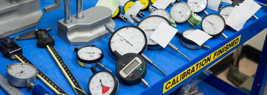 A table for dial gauge calibration. The table has a blue tablecloth and a yellow sign that says "Calibration finished" On top of the table are rows of assorted digital and dial instruments, calipers, and calibration devices with attached certificates of calibration.