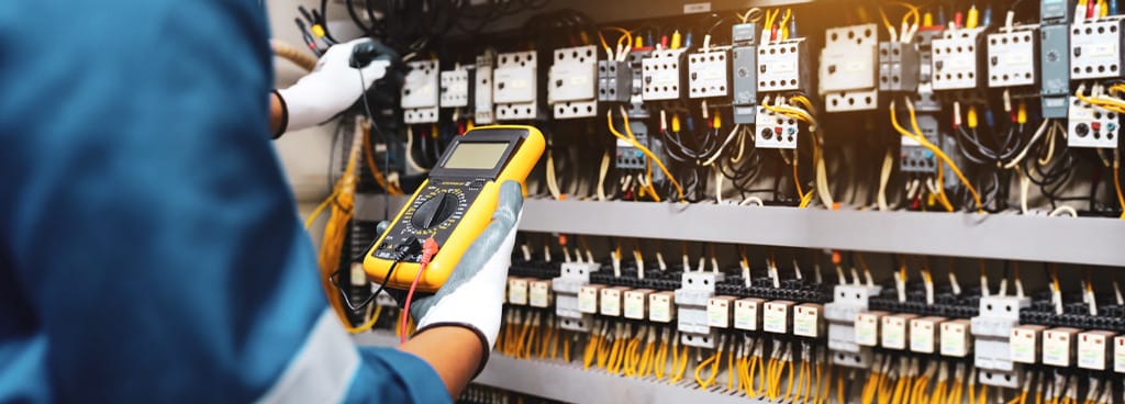 An electrician holds a yellow digital multimeter to read an electric distribution board.