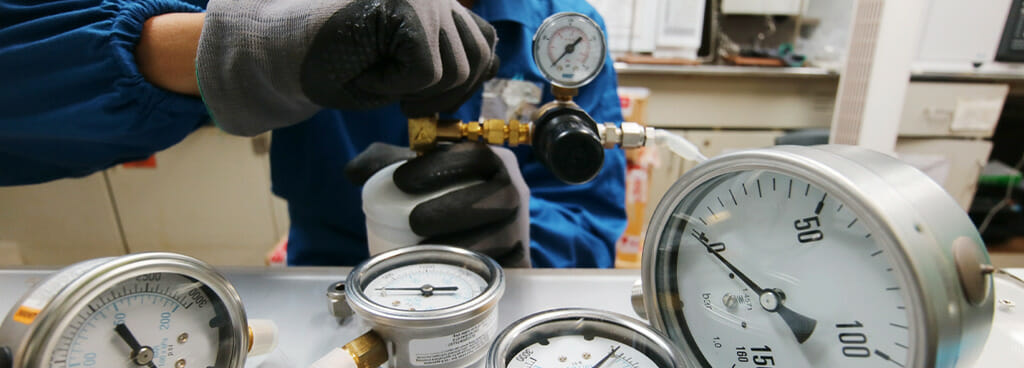 A technician's gloved hands hold a tension/compression gauge. Several other dial gauges lay on a table in the foreground.