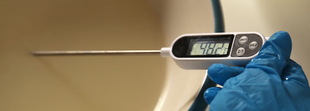 A worker holds a digital thermometer probe in a food processing bowl. The hand wears a blue glove for sanitation.