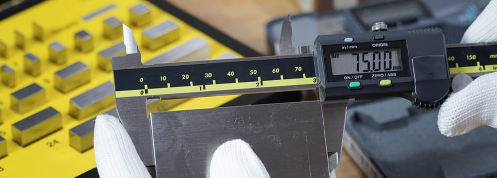 A technician's gloved hands hold a digital caliper as it measures a calibration block. The digital readout says 75.00 mm.