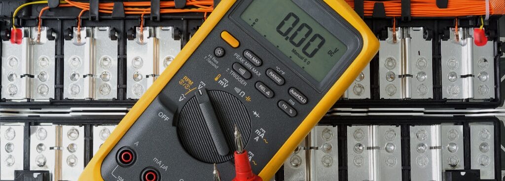A yellow and grey digital multimeter (DMM) rests on top of a module of lithium ion batteries. The multimeter's red and black cables are visible at the bottom of the image.