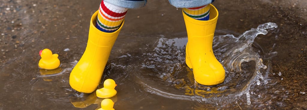 A small child in yellow rain boots jumps in a puddle.
