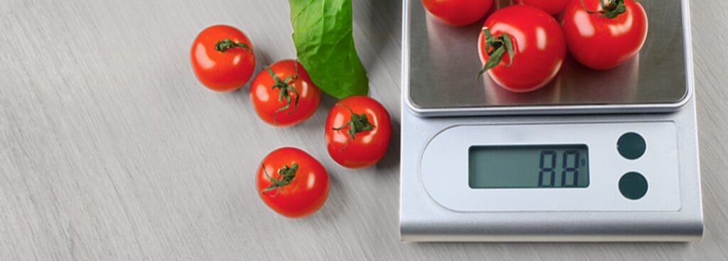 A digital kitchen scale weighs six ripe tomatoes and displays a measurement of 88 grams. Beside the scale are more tomatoes and their leaves.