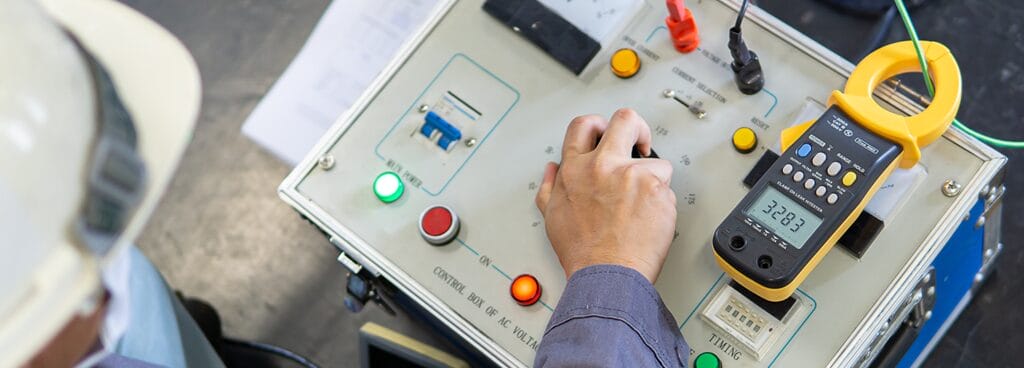 A technician wearing a white hard hat turns the dial for a high voltage generator for dielectric breakdown testing. More electrical controls and lights surround the dial. A yellow and grey handheld meter is installed on top of the machine.