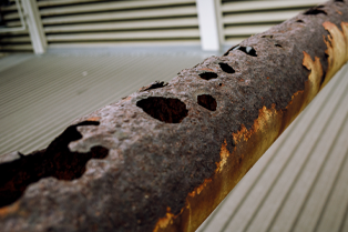 A corroded pipeline. The pipe has multiple holes across the top, with a thick layer of rust surrounding the holes.