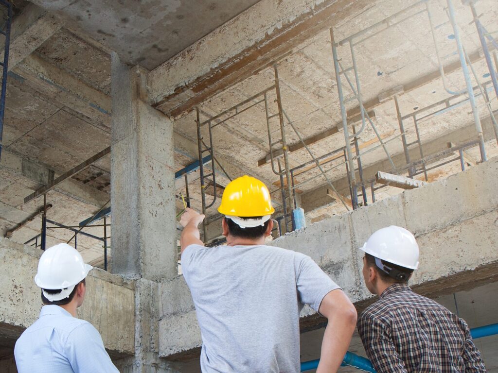 Three structural engineers in a building under construction. One man points to a concrete pillar.