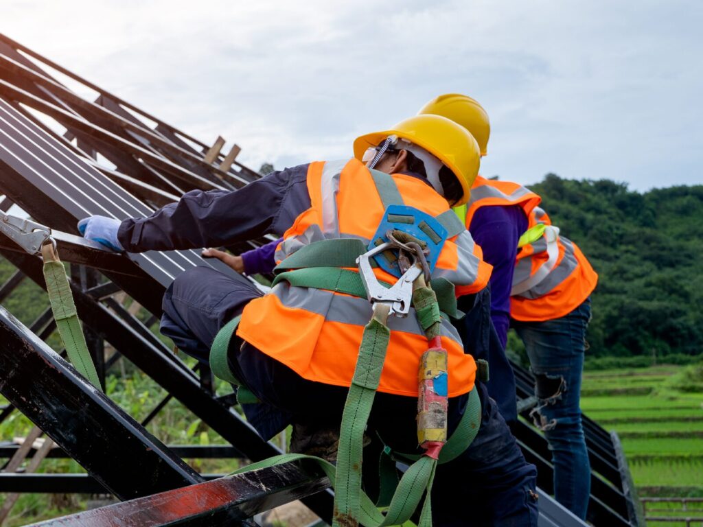 Two construction workers on the steel beams forming a building's structure. They wear harnesses and lanyard to protect them from falls and injuries.