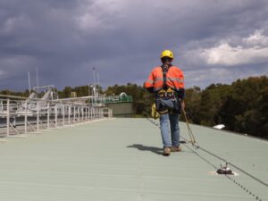 A worker crosses a flat rooftop while. He wears a protective hard hat and harness attached to the roof anchor system via lanyard.