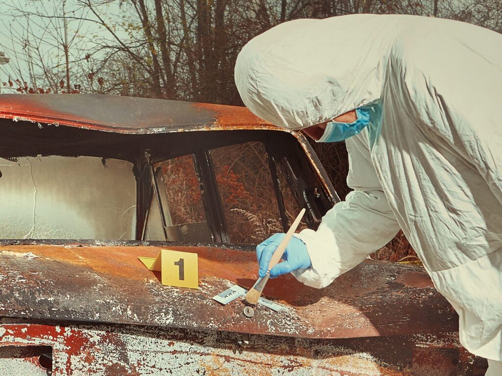 A forensic investigator brushes powder on the keyhole of a burned automobile. An exhibit marker and ruler are placed near the keyhole.
